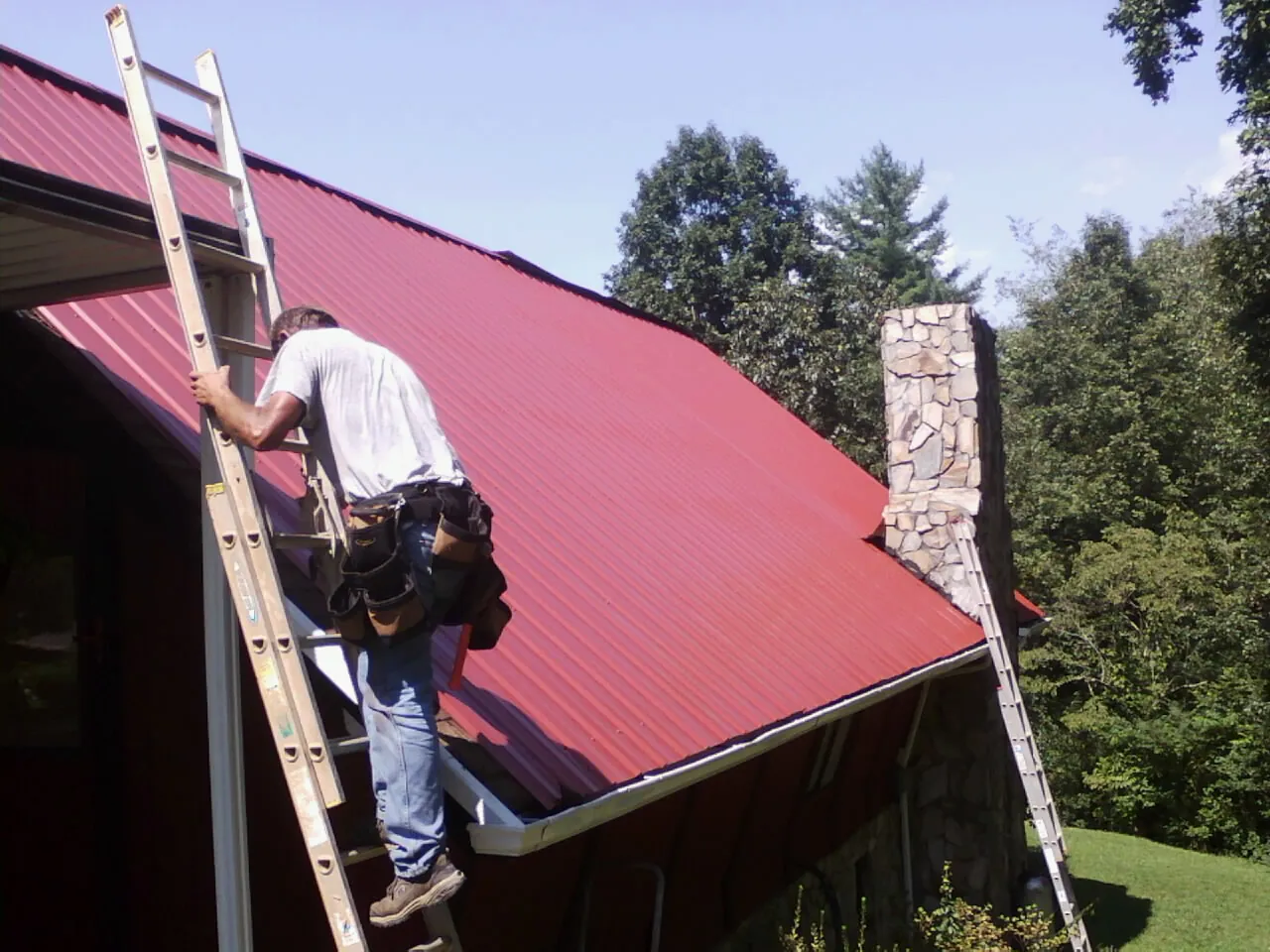 Roofer on a ladder beside a red metal roof and stone chimney — Soffit & Fascia Repair by Roofing Brothers in Percival